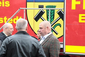 Drei Männer in Anzügen stehen vor einem roten Feuerwehrfahrzeug mit Wappen.