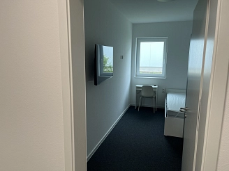 Heller Raum mit Einzelbett, Schreibtisch, Stuhl und Fernseher an der Wand. Ein Fenster bietet Blick auf eine grüne Landschaft.