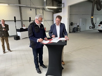 Zwei Männer unterzeichnen Dokumente an einem Tisch in einem hellen, industriell gestalteten Raum.